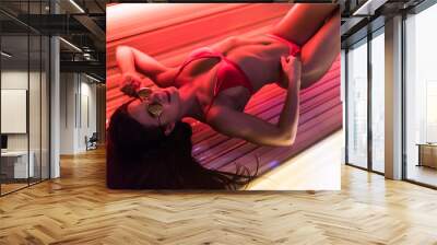 Slim sexy girl in a swimsuit and sunglasses lies in a solarium and tan under the ultraviolet rays Wall mural